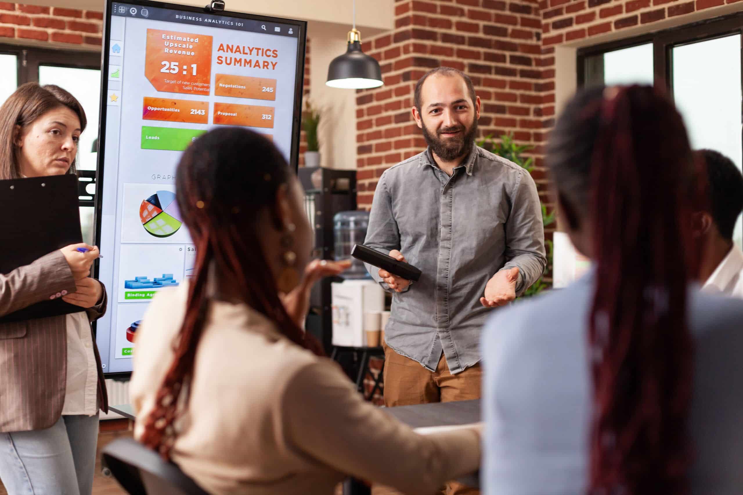 Man Conducting Franchisee Training Programs In An Office Setting, Standing In Front Of A Board And Addressing A Small Audience.